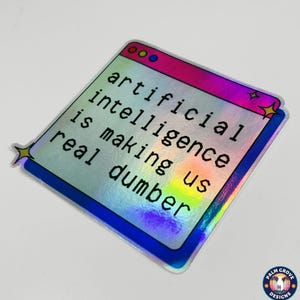 May include: Holographic sticker with the text "artificial intelligence is making us real dumber." The sticker has a window-like design with a pink and blue border and star accents. The Palm Grove Designs logo is in the bottom right corner.