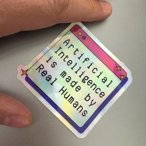 May include: A holographic sticker with the text "Artificial Intelligence is made by Real Humans" in black. The sticker has a window-like design with pink, blue, and yellow accents. The sticker is held by a hand.
