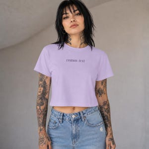 May include: A light purple short-sleeved crop top featuring the words custom text printed in a dark, gothic-style font across the center of the chest. The garment has a crew neckline and a straight hemline that sits above the waistline.