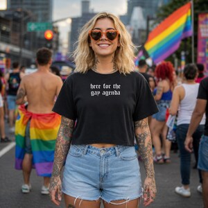 May include: Black crop top with the text "here for the gay agenda" in white gothic font. The person is wearing light blue denim shorts and orange heart-shaped sunglasses. A rainbow flag is visible in the background.