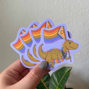 Pode incluir: Uma pilha de adesivos com um dinossauro de desenho animado segurando uma bandeira com listras de arco-íris e um círculo. O dinossauro é marrom com uma gola e pulseiras de arco-íris. Os adesivos têm uma borda roxa clara.