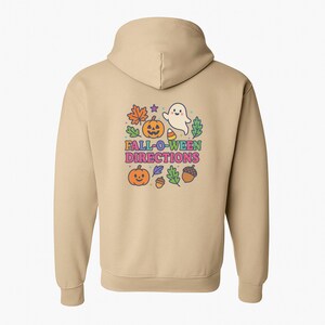 Pode incluir: Um moletom bege com um gráfico colorido nas costas. O gráfico inclui uma abóbora sorridente, um fantasma amigável, folhas de outono e o texto "FALL-O-WEEN DIRECTIONS". O design é em estilo cartoon.