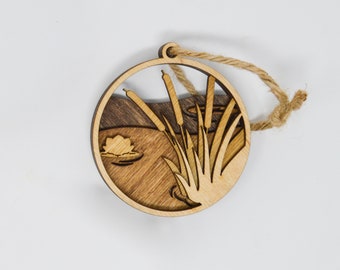 Puede incluir: Un adorno de madera con forma de círculo con un acabado de madera de grano marrón y beige. El adorno presenta un diseño recortado de espadañas y un nenúfar con una flor de nenúfar. El adorno tiene una cuerda de yute para colgar.