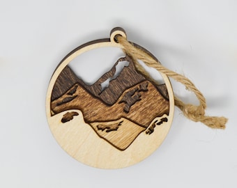 May include: A wooden ornament shaped like a mountain range with a brown and light brown woodgrain pattern. The ornament has a twine string for hanging.