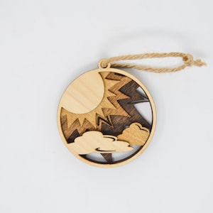 May include: A wooden ornament with a sun, clouds, and lightning bolt design. The ornament is brown and natural wood color and has a twine hanger.