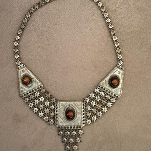 May include: Silver-toned statement necklace with three brown gemstone accents. The necklace features a geometric design with intricate detailing and a chain-link style. The central pendant has a square shape, and the side pendants are triangular.