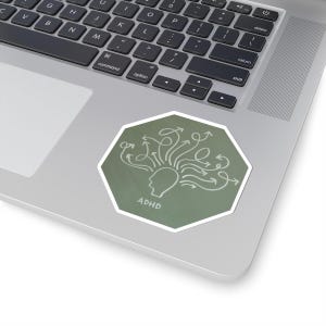 May include: A green, octagonal sticker with a white border. The sticker features a white illustration of a head with arrows emanating from it, and the word "ADHD" below the head. The sticker is placed on a laptop.