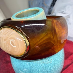May include: A wide, translucent brown tortoiseshell-patterned bangle bracelet featuring a large, oval-shaped, pale peach-colored cabochon set within a polished gold-tone metal frame. The bracelet has a smooth, glossy finish and a rounded profile.