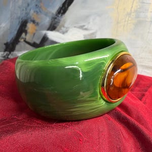 May include: A wide, polished green bangle bracelet featuring a marbled, swirling pattern throughout the material. The bracelet is accented with a single, large, oval-shaped amber-colored cabochon stone set within a smooth, shiny gold-tone metal bezel.