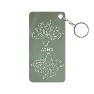 Può includere: Un portachiavi rettangolare verde salvia con catena e anello argentati. Il portachiavi presenta un disegno a tratto bianco di una testa con frecce e la parola "ADHD". Sotto c'è una seconda testa con frecce.