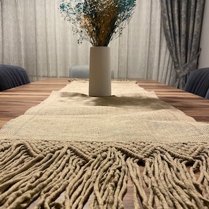 May include: A beige table runner with long fringe on a wooden table. A white vase with dried flowers in shades of blue and beige is in the center of the table.