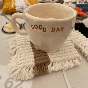 Może przedstawiać: Biały kubek ceramiczny z napisem "GOOD DAY" w pomarańczowych literach. Kubek stoi na białym i brązowym plecionym podkładce.