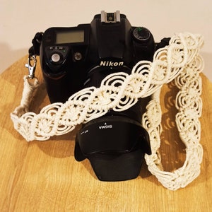 May include: A black Nikon camera with a white crocheted camera strap. The camera lens is a Sigma lens.