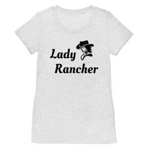 May include: Light gray t-shirt with the words "Lady Rancher" in a script font, accompanied by a black silhouette of a woman wearing a cowboy hat. The shirt has a scoop neck and short sleeves.