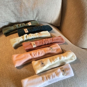 Op de afbeelding: Een set van acht zachte, pluche hoofdbanden in verschillende kleuren, waaronder wit, crème, roze en groen. Elke hoofdband heeft een klein label met de tekst "JoJo" erop.