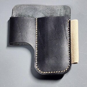 Pode incluir: Estojo de couro preto com costura bege e uma faixa elástica bege. O estojo tem um design curvo e uma aba. O couro parece texturizado. O estojo provavelmente foi projetado para guardar um item pequeno.