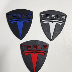 May include: Three black Tesla emblem patches with a shield shape. Each patch features the word "TESLA" above a "T" logo. The logos are colored blue, white, and red.