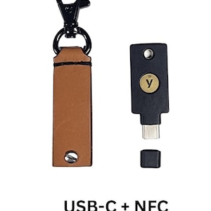 Może przedstawiać: Czarny klucz bezpieczeństwa USB-C i NFC z odpinaną nasadką, obok brązowego skórzanego breloka z czarnym metalowym zapięciem. Klucz ma złoty przycisk z białym symbolem "y". Tekst "USB-C + NFC" znajduje się na dole.
