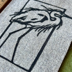 May include: A gray felt square featuring a black graphic of a bird within a rectangular frame. The bird is depicted in a minimalist style, with long legs and a curved neck. The felt has a textured appearance.