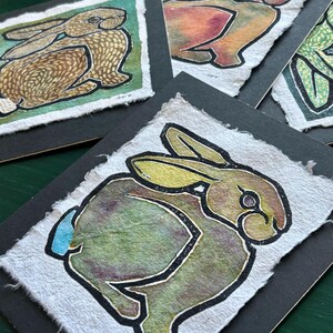 May include: A collection of handmade art cards featuring watercolor rabbit illustrations. Each card showcases a unique rabbit design with a black outline, set against a textured paper background. The rabbits are depicted in various colors, including brown, orange, and green.