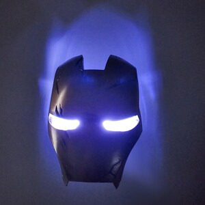 May include: A black and silver Iron Man mask with glowing white eyes. The mask is lit from behind, casting a blue glow on the wall.