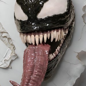 May include: A detailed sculpture of the Venom character's head, featuring a glossy black exterior, white eyes, and a long, pink tongue. The mouth is open, revealing sharp, white teeth. The sculpture is mounted on a textured, cracked wall.