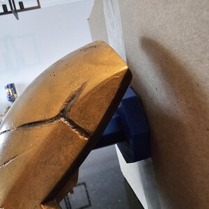 May include: A gold-coloured helmet with a textured surface is being held in place by a blue clamp. The helmet is attached to a piece of cardboard.