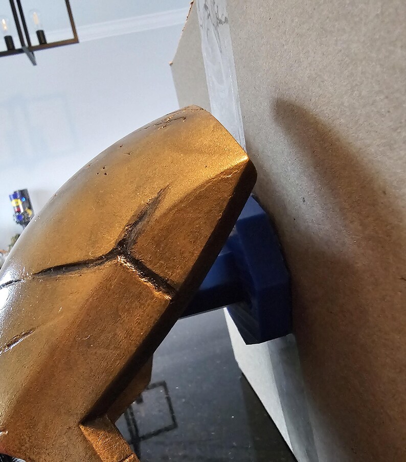 May include: A gold-coloured helmet with a textured surface is being held in place by a blue clamp. The helmet is attached to a piece of cardboard.