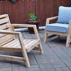 K&ouml;nnte beinhalten: Zwei Holzst&uuml;hle mit Lattenr&uuml;cken und Armlehnen. Ein Stuhl hat ein graues Kissen. Die St&uuml;hle stehen auf einer Terrasse aus Ziegeln.