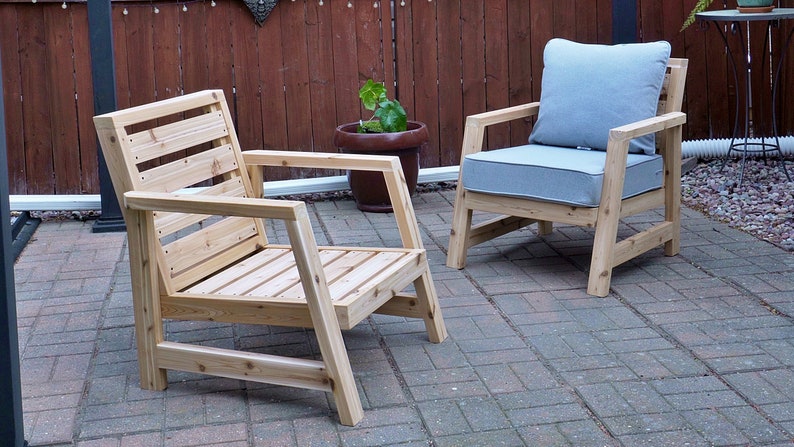 K&ouml;nnte beinhalten: Zwei Holzst&uuml;hle mit Lattenr&uuml;cken und Armlehnen. Ein Stuhl hat ein graues Kissen. Die St&uuml;hle stehen auf einer Terrasse aus Ziegeln.