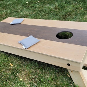 May include: A wooden cornhole game set with two gray bean bags. The game board has a brown and beige striped design with a hole in the center.