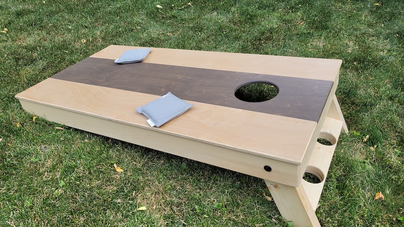 May include: A wooden cornhole game set with two gray bean bags. The game board has a brown and beige striped design with a hole in the center.