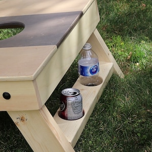 May include: A wooden cornhole game with a brown and beige playing surface and a built-in drink holder. A clear plastic water bottle and a brown aluminum can are in the drink holder.