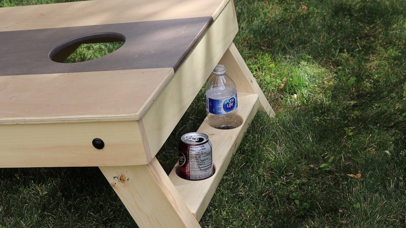 May include: A wooden cornhole game with a brown and beige playing surface and a built-in drink holder. A clear plastic water bottle and a brown aluminum can are in the drink holder.