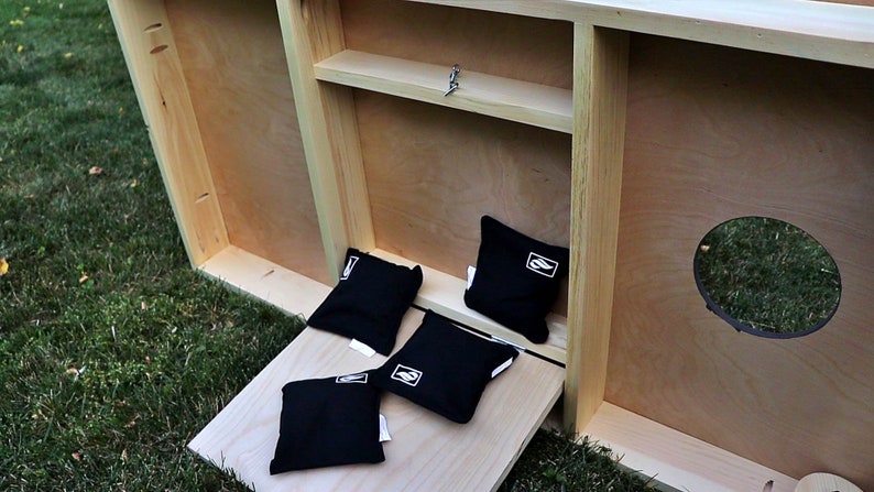 May include: A wooden cornhole game with a storage compartment and a hinged door. The game is set up on a grassy surface with four black bean bags.