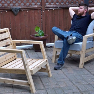 K&ouml;nnte beinhalten: Zwei Holzst&uuml;hle mit Lattenr&uuml;cken und Armlehnen. Ein Stuhl ist leer, auf dem anderen sitzt eine Person. Die St&uuml;hle stehen auf einer Terrasse mit einem Ziegelsteinboden.