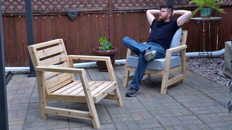 K&ouml;nnte beinhalten: Zwei Holzst&uuml;hle mit Lattenr&uuml;cken und Armlehnen. Ein Stuhl ist leer, auf dem anderen sitzt eine Person. Die St&uuml;hle stehen auf einer Terrasse mit einem Ziegelsteinboden.