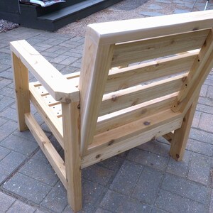 K&ouml;nnte beinhalten: Ein Holzstuhl mit einer Lattenlehne und Armlehnen. Der Stuhl ist aus unbehandeltem Holz und hat eine nat&uuml;rliche Holzfarbe.