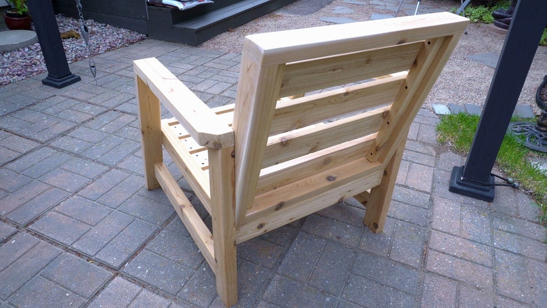 K&ouml;nnte beinhalten: Ein Holzstuhl mit einer Lattenlehne und Armlehnen. Der Stuhl ist aus unbehandeltem Holz und hat eine nat&uuml;rliche Holzfarbe.