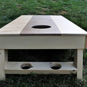 May include: A wooden cornhole game with a light brown top and a white base. The top has a circular hole in the center and two smaller holes on the bottom for holding bean bags.