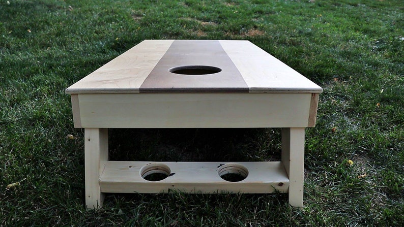 May include: A wooden cornhole game with a light brown top and a white base. The top has a circular hole in the center and two smaller holes on the bottom for holding bean bags.