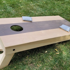 May include: A wooden cornhole game with a brown and natural wood finish. The game has two bean bags, one on each side of the board. The board is set up on grass.