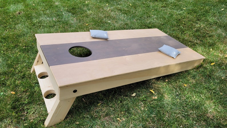 May include: A wooden cornhole game with a brown and natural wood finish. The game has two bean bags, one on each side of the board. The board is set up on grass.