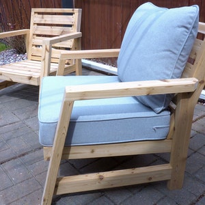 K&ouml;nnte beinhalten: Zwei Holzst&uuml;hle mit hellgrauen Kissen. Die St&uuml;hle haben ein schlichtes, modernes Design mit einer Lattenr&uuml;cklehne und breiten Armlehnen. Die St&uuml;hle sind aus Naturholz gefertigt und haben einen rustikalen Look.