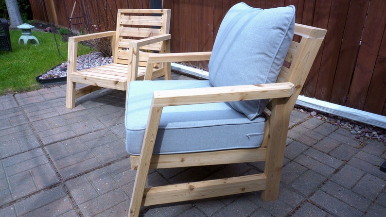 K&ouml;nnte beinhalten: Zwei Holzst&uuml;hle mit hellgrauen Kissen. Die St&uuml;hle haben ein schlichtes, modernes Design mit einer Lattenr&uuml;cklehne und breiten Armlehnen. Die St&uuml;hle sind aus Naturholz gefertigt und haben einen rustikalen Look.