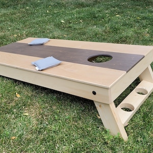May include: A wooden bean bag toss game with a brown and beige playing surface. The game has two bean bags and a hole in the center of the playing surface.