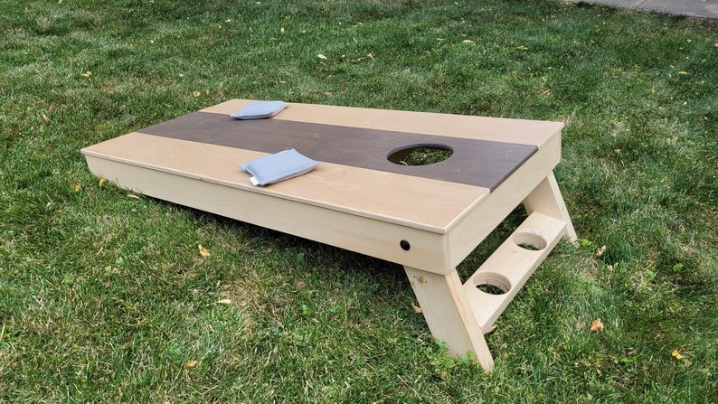 May include: A wooden bean bag toss game with a brown and beige playing surface. The game has two bean bags and a hole in the center of the playing surface.