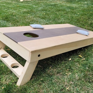 May include: A wooden cornhole game with a brown and beige playing surface. The game has two bean bags on the surface and two cup holders on the side.