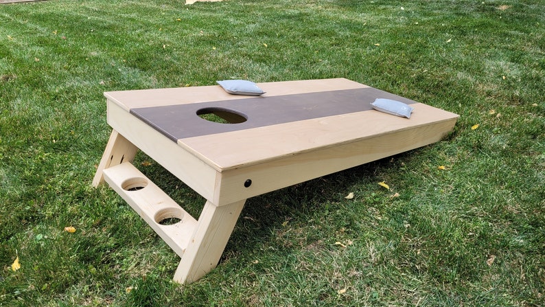 May include: A wooden cornhole game with a brown and beige playing surface. The game has two bean bags on the surface and two cup holders on the side.