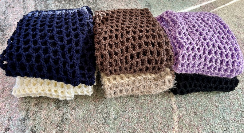 May include: A collection of five folded crochet items featuring an open, grid-like stitch pattern. The colors include dark navy blue, cream, brown, beige, lavender, and black, all stacked in a neat arrangement to display the varied yarn textures.